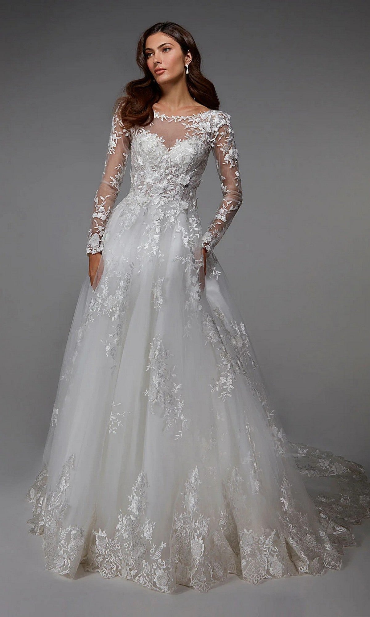 A woman wears the Alyce 7046, a long-sleeved, floor-length white dress with floral embroidery and a sheer neckline, standing against a plain gray background.